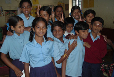 A group of children smiling and posing together for a photo, standing close to each other, some making playful gestures, in a bright and cheerful setting.