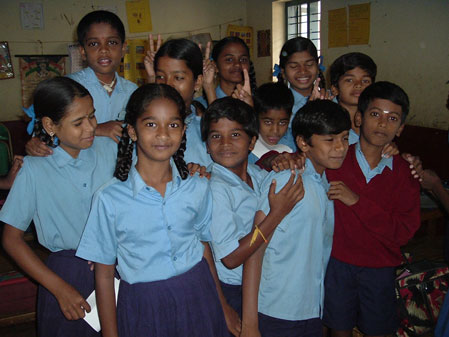 A group of children smiling and posing together for a photo, standing close to each other, some making playful gestures, in a bright and cheerful setting.