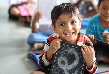 A boy holding a slate and smiling brightly, looking happy and proud in a classroom setting.